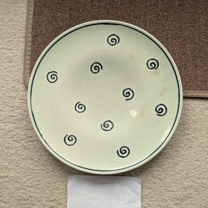 Peut inclure: Une assiette ronde en céramique blanc cassé, bordée d'un liseré bleu foncé et ornée de motifs en spirale noirs espacés. L'assiette est posée sur une surface beige, avec un tapis marron en arrière-plan. Un papier blanc est au premier plan.