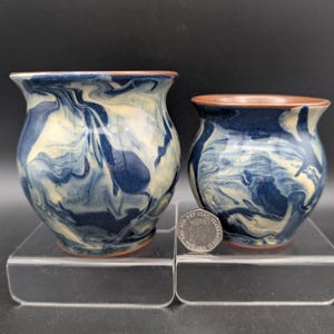 May include: Two ceramic vases with a marbled blue and cream design. The vases have a rounded shape with a flared rim and a brown rim. The vases are displayed on a clear acrylic surface.