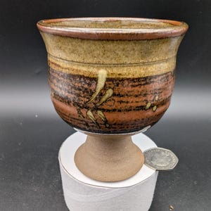Może przedstawiać: Ręcznie robiony ceramiczny kielich z szerokim, lekko rozszerzonym brzegiem. Kielich ma brązowo-beżową kolorystykę z dekoracyjnym kwiatowym wzorem. Kielich ma około 10 cm wysokości.