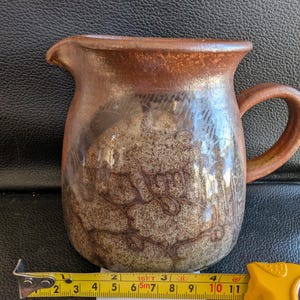 May include: A brown and white ceramic pitcher with a speckled glaze. The pitcher has a rounded body and a handle.