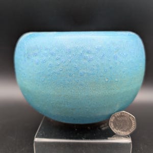 Op de afbeelding: Een kleine, ronde, turquoise keramische kom met een textuuroppervlak. De kom wordt gepresenteerd op een heldere acrylstandaard. Een munt ligt naast de kom ter vergelijking van de grootte. De achtergrond is zwart.