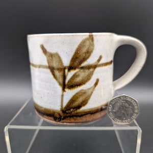 May include: A ceramic mug with a white base and a brown leaf design. The mug has a curved handle and a brown band around the top and bottom. The mug is approximately 7.6 cm tall.