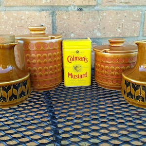 Può includere: Un set di quattro contenitori per condimenti in ceramica marrone e arancione con un motivo floreale. I contenitori sono su un tavolo in metallo nero. Una lattina gialla di senape Colman's è al centro del tavolo.