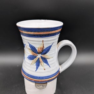 Può includere: Tazza in ceramica bianca con un motivo floreale blu e marrone. La tazza ha una base larga e un manico.