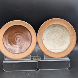 Könnte beinhalten: Zwei handgefertigte Keramikplatten. Eine Platte ist braun mit einem Spiralmuster, die andere ist weiß mit braunen Sprenkeln. Beide Platten haben einen hellbraunen Rand. Die Platten werden auf einem transparenten Acrylständer ausgestellt.
