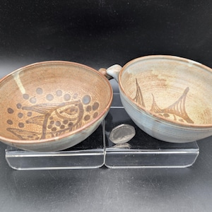 May include: Two ceramic bowls with handles, each featuring a hand-painted brown fish design on the interior. The bowls have a speckled tan and grey glaze. A small silver-coloured coin is placed between the bowls for scale on the clear base.