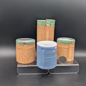 May include: Four ceramic vessels of varying heights and colors. Two are cylindrical with a textured, basket-weave pattern in tan, with green and brown rims. One is blue with a grid pattern, and the tallest is tan with a green rim and button closure.