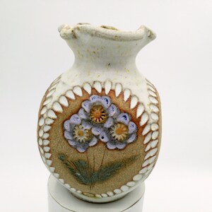 May include: A small, white ceramic vase with a brown and white floral design. The vase has a round body and a narrow neck. The flowers are purple with yellow centres.