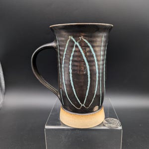 May include: A black ceramic mug with a handle, featuring a light blue vertical design. The mug has a textured surface and a tan base. The mug is displayed on a clear stand against a black background.