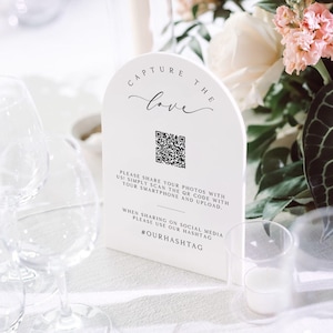 Op de afbeelding: Een wit bord met een afgeronde boog bovenaan waarop in zwarte letters "Capture the love" staat. Onder de tekst staat een zwart-witte QR-code met instructies om de code te scannen met een smartphone en foto's te uploaden. De tekst bevat ook de hashtag #OURHASHTAG.