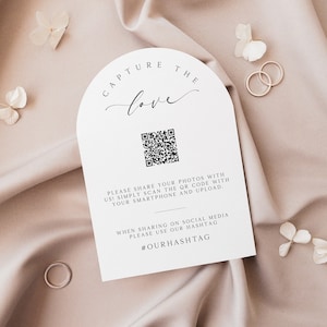 Puede incluir: Cartel blanco arqueado con el texto "CAPTURE THE love" y un código QR. El cartel incluye instrucciones para compartir fotos con un smartphone y un hashtag. También hay dos anillos dorados y flores blancas.