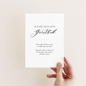 May include: A white card with black text that reads "PLEASE SIGN OUR Guestbook". The text continues, "Pick a page and leave a note or simply sign your name. This book will be a memory of all the guests who came to celebrate with us."