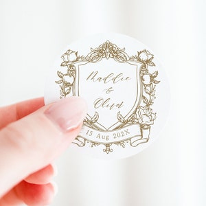 May include: A white round sticker with a gold design of a crest with the text "Maddie & Oliver" and the date "15 Aug 202X".