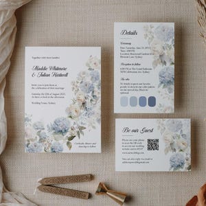 May include: Wedding stationery set with blue and white floral arrangements. The set includes an invitation, details card, and a guest card with a QR code. The invitation displays names, date, and location details.