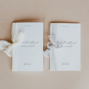 Könnte beinhalten: Zwei weiße Hochzeitszeremonie-Programme mit einem Band um jedes gebunden. Die Programme sind mit "to the wedding of Cassie & Thomas" und "to the wedding of . & Thomas" betitelt. Die Programme sind datiert auf den 1. August 2023, Hyde Park, Sydney.