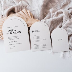Op de afbeelding: Een set van drie huwelijksuitnodigingskaarten met een modern boogontwerp. De kaarten zijn gedrukt op crèmekleurig papier met zwarte tekst. De hoofkaart toont de namen Amelia en Benjamin en de datum van de bruiloft. De tweede kaart bevat de details van de ceremonie en de receptie. De derde kaart heeft de initialen A en B.