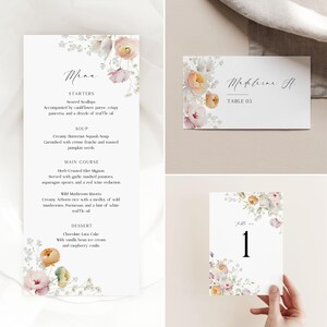 Pode incluir: Um conjunto de papelaria de casamento elegante com menu, cartão de lugar e número da mesa. Os designs têm um fundo branco com detalhes florais em tons de rosa claro, pêssego e verde. O menu lista entradas, sopa, prato principal e sobremesa.