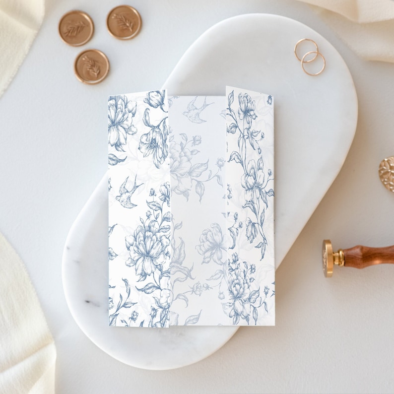 May include: A folded white card with a blue floral pattern and small birds. The card is on a white marble surface.