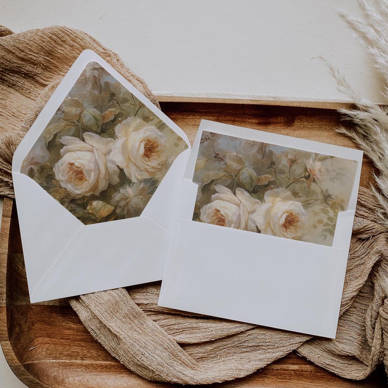 May include: Two white envelopes with a floral design on the front. The design features white roses and green leaves on a light brown background.