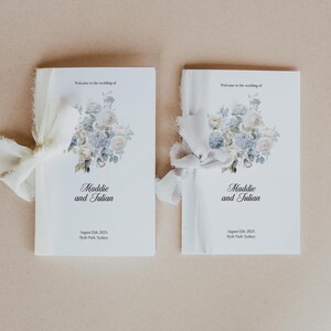 May include: Two white wedding programs tied with ivory ribbons. Each program features a floral design in pastel colours and the names "Maddie and Julian." The date and location, August 12th, 2021, Hyde Park, Sydney, are also printed.