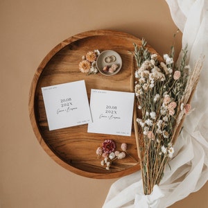 May include: Two white save the date cards with the text 'Save Our Date' and '20.08.202X' in black text. The cards are on a wooden tray with dried flowers and two gold rings on a beige surface.