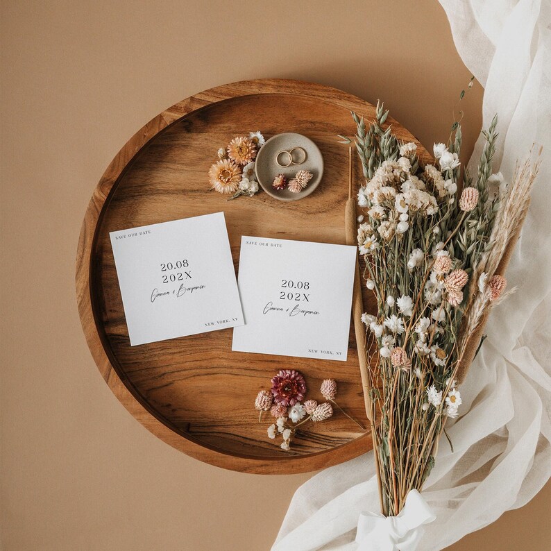 May include: Two white save the date cards with the text 'Save Our Date' and '20.08.202X' in black text. The cards are on a wooden tray with dried flowers and two gold rings on a beige surface.