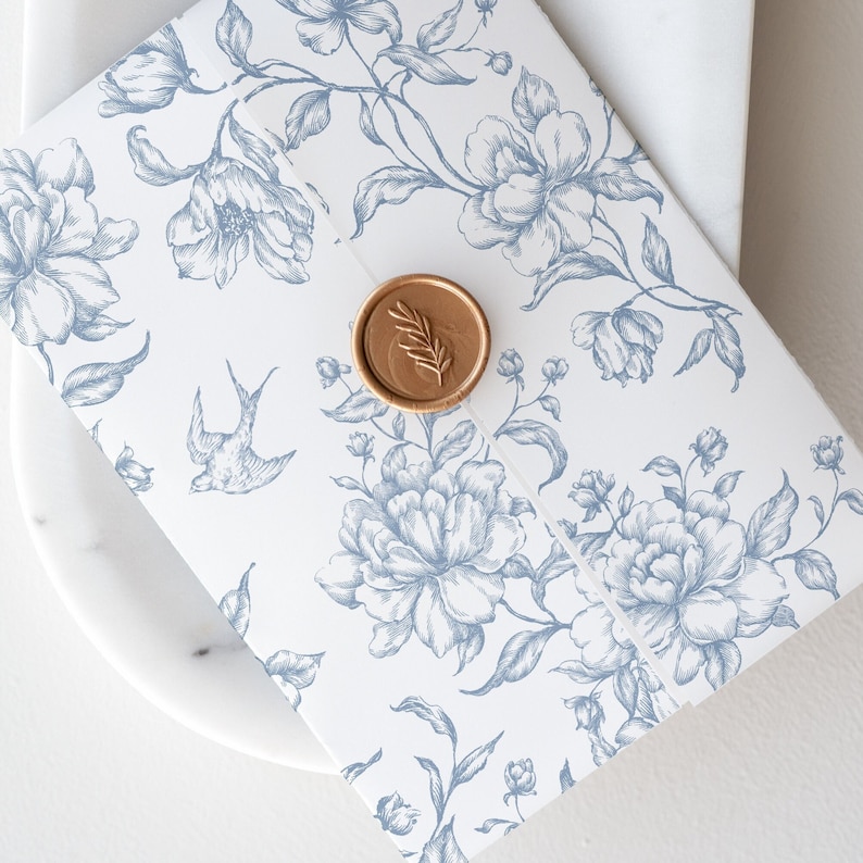 May include: A white envelope with a blue floral pattern and a gold wax seal. The envelope is on a white marble surface.