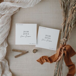 May include: Two white save the date cards with black text. The text reads 'Save Our Date', '20.08 20XX', 'Cowen & Bapon', and 'NEW YORK, NY'. The cards are on a beige surface with a brown ribbon and dried grass. 