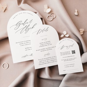 May include: Elegant wedding invitation suite with arched cards. The main card features the names "Giulia & Elijah" in cursive script. Details card includes ceremony information. RSVP card with QR code. Two gold rings and floral accents.