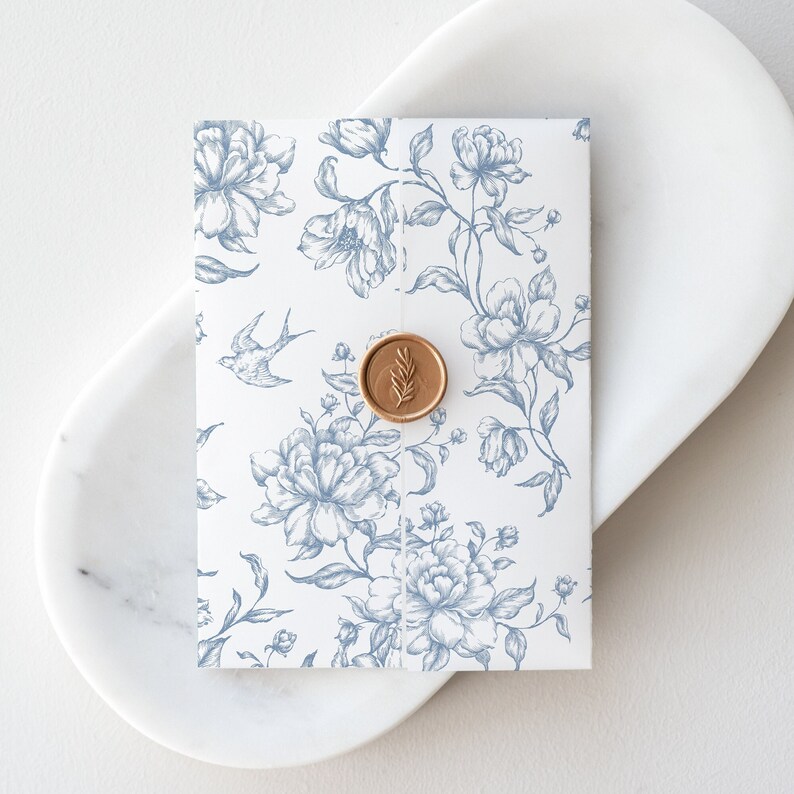 May include: A white envelope with a blue floral pattern and a gold wax seal. The envelope is on a white marble surface.