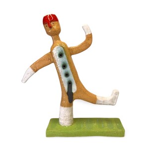 May include: A ceramic figurine of a person wearing a red hat and a blue and white patterned coat. The figurine is standing on a green base and has a white and brown body.