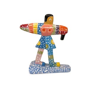 May include: A colorful, handcrafted wooden figurine of a person with outstretched arms. The figure has a black head, orange face with red eyes, and a blue dress with yellow sun designs. The arms are striped with various colors, and the base is blue.