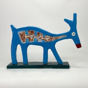 Könnte beinhalten: Eine blaue Keramikskulptur eines stilisierten Hirsches mit braunen und weißen Details. Der Hirsch steht auf einem dunkelgrünen Sockel.