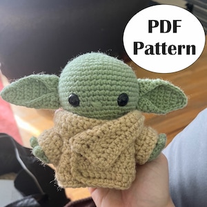 Könnte beinhalten: Ein gehäkeltes grünes und braunes Plüschtier, das einem Charakter aus dem Star Wars-Franchise ähnelt. Das Spielzeug trägt einen braunen Pullover und hat große schwarze Augen. Das Bild enthält den Text "PDF Pattern".