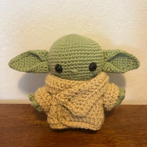 Pu&ograve; includere: Figura all'uncinetto di un personaggio con grandi orecchie, pelle verde e occhi neri. La figura indossa un capo con cappuccio color beige. L'articolo fatto a mano &egrave; esposto su una superficie di legno.