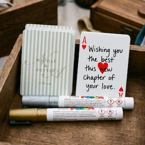 Puede incluir: Un mazo de cartas con un patrón de rayas verde salvia, personalizado con los nombres Nicole & Austin y la fecha 07.24.2026. Junto a él, un as de corazones muestra el texto Wishing you the best this new chapter of your love en negro.