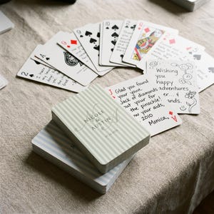 Peut inclure: Un jeu de cartes à dos rayé vert clair et blanc portant le texte Nicole & Austin et la date 07.24.2026. Plusieurs cartes sont étalées, montrant des messages manuscrits, des enseignes noires et rouges, et une dame de carreau.