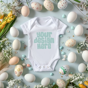 May include: A white baby onesie with the text "your design here" in a light blue colour. The onesie is surrounded by white and patterned Easter eggs, flowers, and sweets.