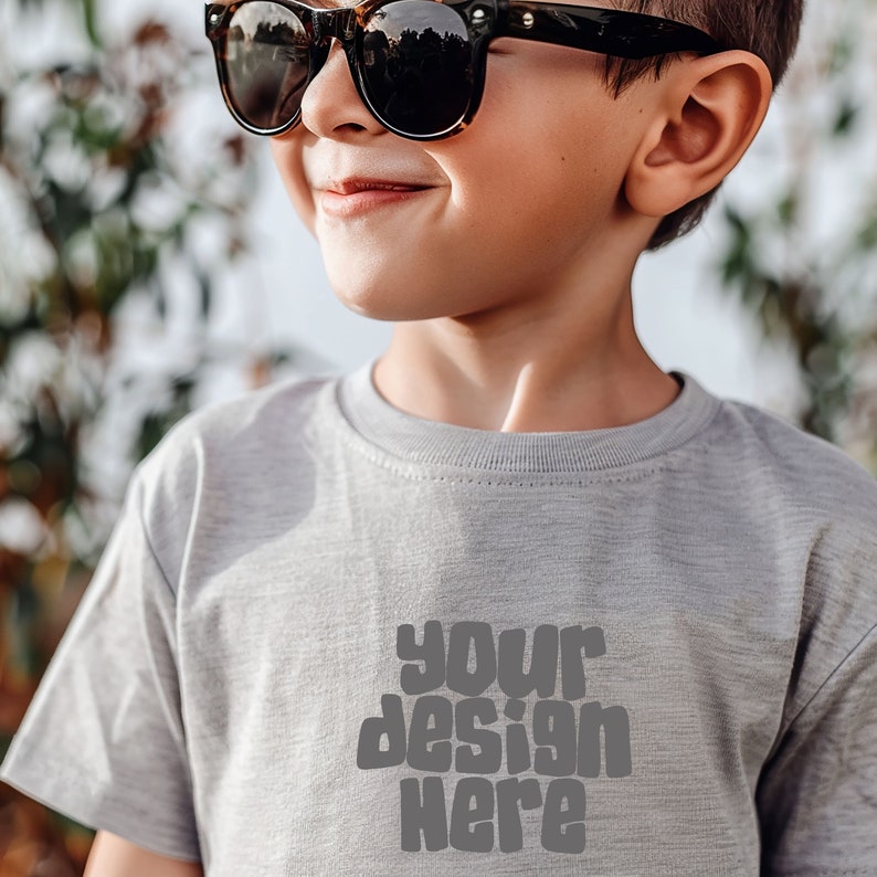 May include: A young boy wearing a gray t-shirt with the text "your design here" printed on the front. He is wearing brown sunglasses and looking to the right.