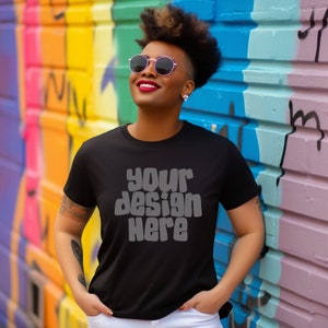 May include: A black t-shirt with the text "your design here" in a retro font. The shirt is being worn by a person with short, curly hair and sunglasses. The person is standing in front of a colorful wall.