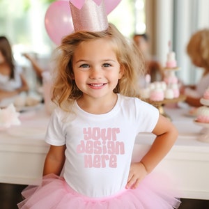 May include: A young girl wearing a white t-shirt with the text "your design here" in pink letters. She is wearing a pink tutu skirt and a pink and silver crown. She is smiling at the camera.