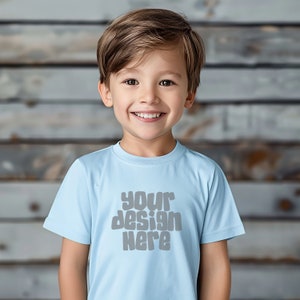 May include: A young boy wearing a light blue t-shirt with the text "your design here" in gray. The boy is smiling and looking at the camera.