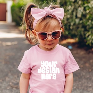 May include: A young child wearing a pink t-shirt with the text "your design here" in white. The child is also wearing pink sunglasses and a pink bow headband.