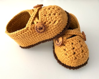 Könnte beinhalten: Ein Paar handgefertigte Baby-Häkel-Schuhe in Senfgelb mit braunen Verzierungen. Die Schuhe haben Holzknöpfe mit einem Kronenmuster. Das Häkelmuster ist sichtbar, vor einem weißen Hintergrund.