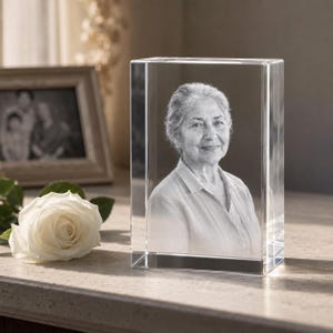 May include: A rectangular clear glass block featuring a laser-etched black and white portrait of an older individual with short hair, wearing a collared shirt. The glass block has sharp, clean edges and a smooth, transparent surface finish.