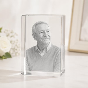 May include: A rectangular clear glass block featuring a laser-etched portrait of an older individual with short hair, wearing a collared shirt under a V-neck sweater. The portrait is rendered in shades of gray and white within the solid glass.