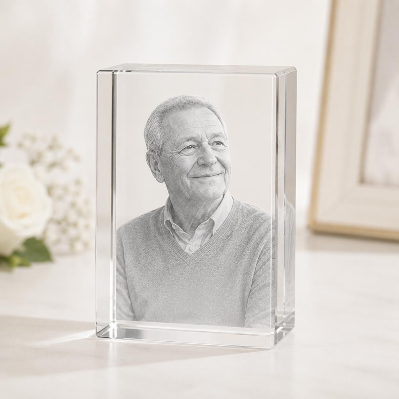 May include: A rectangular clear glass block featuring a laser-etched portrait of an older individual with short hair, wearing a collared shirt under a V-neck sweater. The portrait is rendered in shades of gray and white within the solid glass.