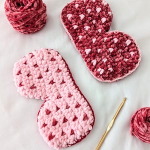 Puede incluir: Primer plano de artículos de ganchillo hechos a mano en tonos rosa y burdeos. Se ven dos objetos con forma de zapatilla, junto con dos ovillos de lana y un ganchillo dorado. Los artículos están hechos con un hilo suave y texturizado.