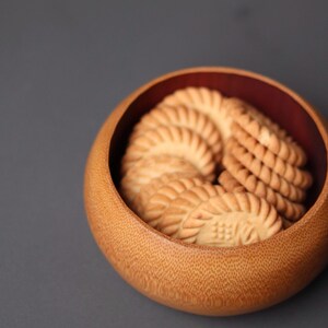 K&ouml;nnte beinhalten: Eine Holzschale mit hellbraunem &Auml;u&szlig;eren und dunkelrotem Innenraum, gef&uuml;llt mit mehreren goldbraunen, gemusterten Keksen. Die Kekse haben ein geriffeltes Design und sind spiralf&ouml;rmig angeordnet.