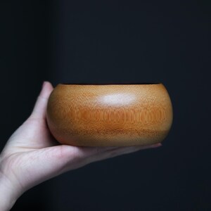 K&ouml;nnte beinhalten: Eine kleine, runde Holzschale mit nat&uuml;rlicher, hellbrauner Oberfl&auml;che. Die Schale wird in einer Hand vor einem dunklen Hintergrund gehalten. Die glatte, geschwungene Form der Schale und die Holzmaserung sind sichtbar.
